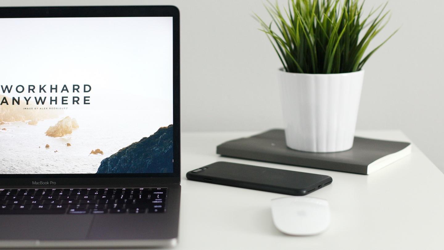 A laptop with the words "work hard anywhere" on it's screen on a minimal work from home set-up with a plant and notebook